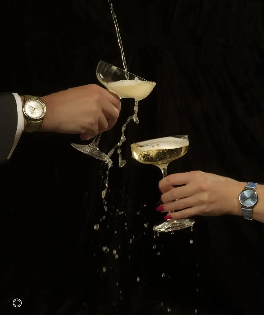 Close up view of champagne being poured into two wide champagne glasses held by different hands against a black background.